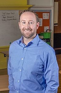 John Dues, a white man smiling and wearing a blue shirt , standing in a classroom