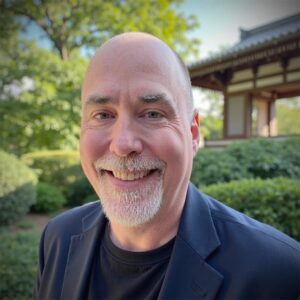 Michael J. Cousineau, a white man with closely cropped hair and a trim white beard, standing outside by a Japanese-style garden and pavilion.