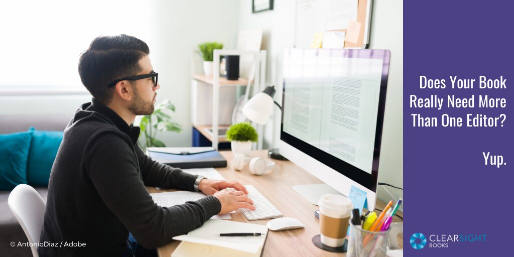 Young white man with dark hair and beard, wearing glasses and a dark shirt, sitting at a computer editing a book.