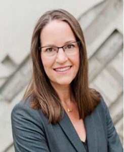 Marci Shepard, a middle aged white woman with shoulder-length brown hair and glasses wearing a greenish suit jacket and smiling at the viewer.