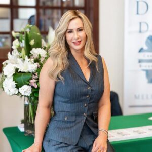 Melissa Hull, a white woman with long blond hair, wearing a navy pinstriped suit vest and pants and leaning against a table with flowers on it. Her book is visible on the table and she is smiling directly at the camera.