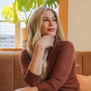 Melissa Hull, a white woman with long blond hair, wearing a rust-colored shirt and sitting on a brown sofa with a plant in the background; her hand is on her chin and she is looking into the distance with a slight smile on her face.