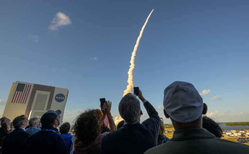 People watching and taking photos of the Artemis II launch. A bright blue sky with a tiny rocket shooting up with a big cloud following it. A NASA building with a giant American flag is in the background at left.