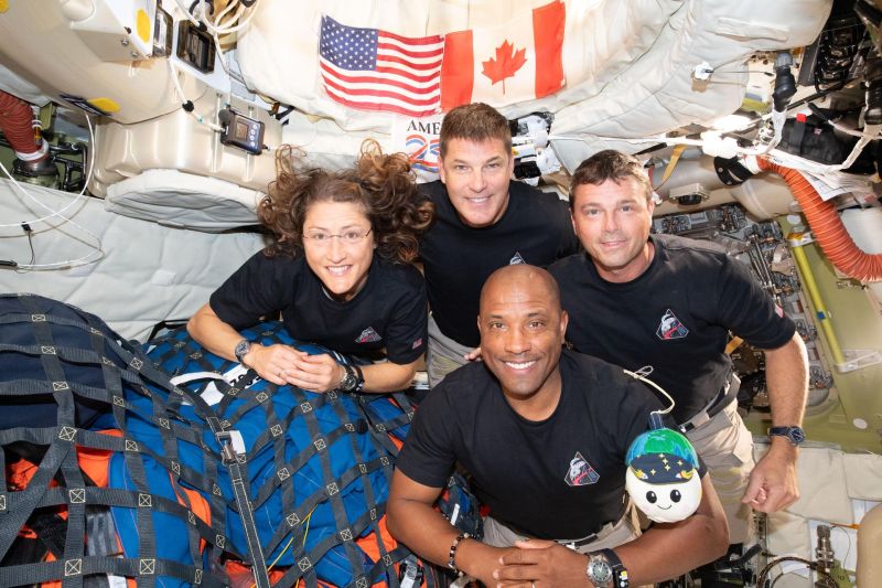 The four crew members wearing black t-shirts in their cabin. Koch is a white woman with longer curly hard; Hansen is a white man with short brown hair; Wiseman is a white man with short brown hair; Glover is a black man with a shaved head. They are in zero gravity (shown by Koch's floating hair) and have their toy, Rise, with them. The American and Canadian flags are in the background and everything is secured to not float away. They are all smiling.