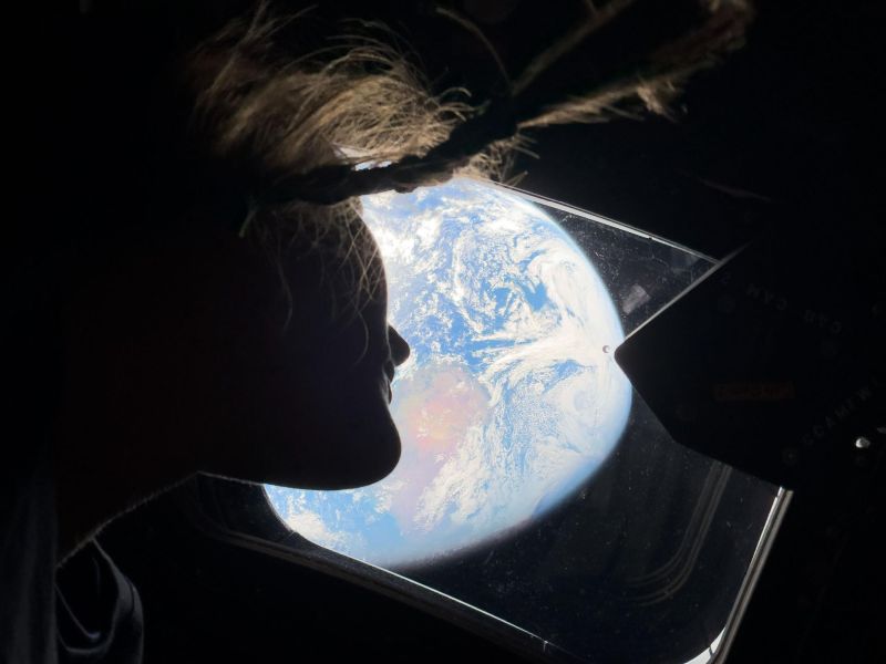 Christina Koch looking through the window at the Earth in the distance. The camera is angled up so you see Koch's chin from bottom and her longer hair is floating around. The Earth is bright so everything else seems dark.