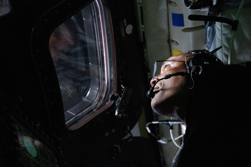 Victor Glover is looking to the left out the window. The camera angle is tilted up at his chin, and the left side of his face is illuminated, presumably from what he is viewing out the window.