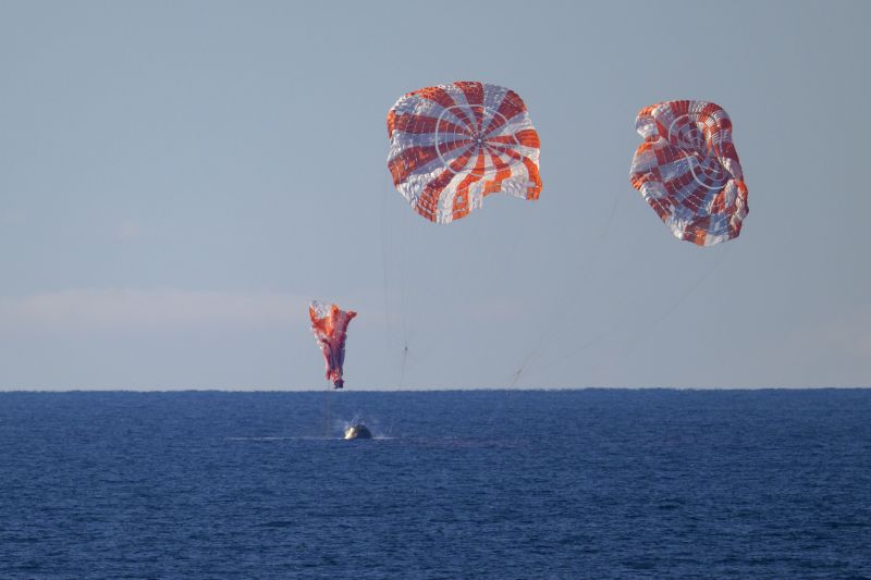 Three orange and white striped parachutes collapsing as the reentry capsule lands in the blue ocean.