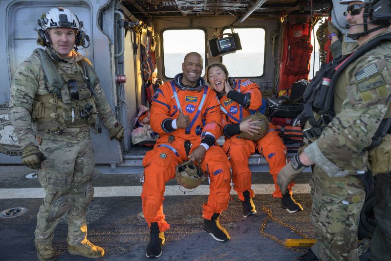 Glover and Koch in their bright orange space suits sitting on a helicopter with military support staff around them. They are smiling big and Glover is giving a thumbs-up.