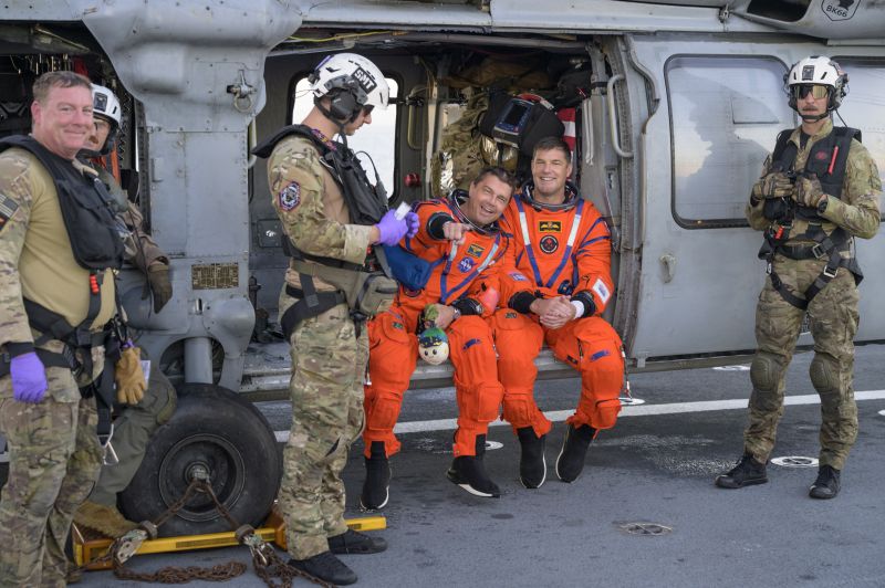 Wiseman and Hansen in their bright orange space suits sitting on a helicopter with military support staff around them. They are smiling big and Wiseman is pointing at the camera.