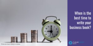 Three stacks of coins—short, medium, tall—next to a pastel green alarm clock. Text: When is the best time to write your book?